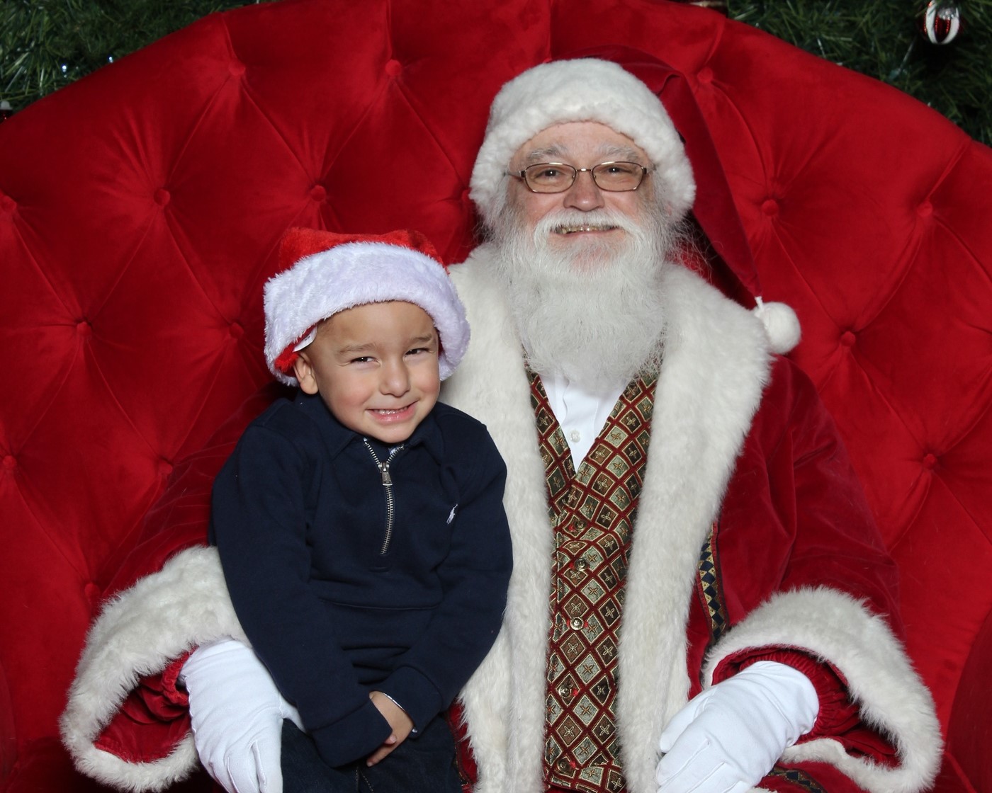 A child in a Santa hat sits on Santa Claus’s lap, both smiling against a backdrop of plush red upholstery.
